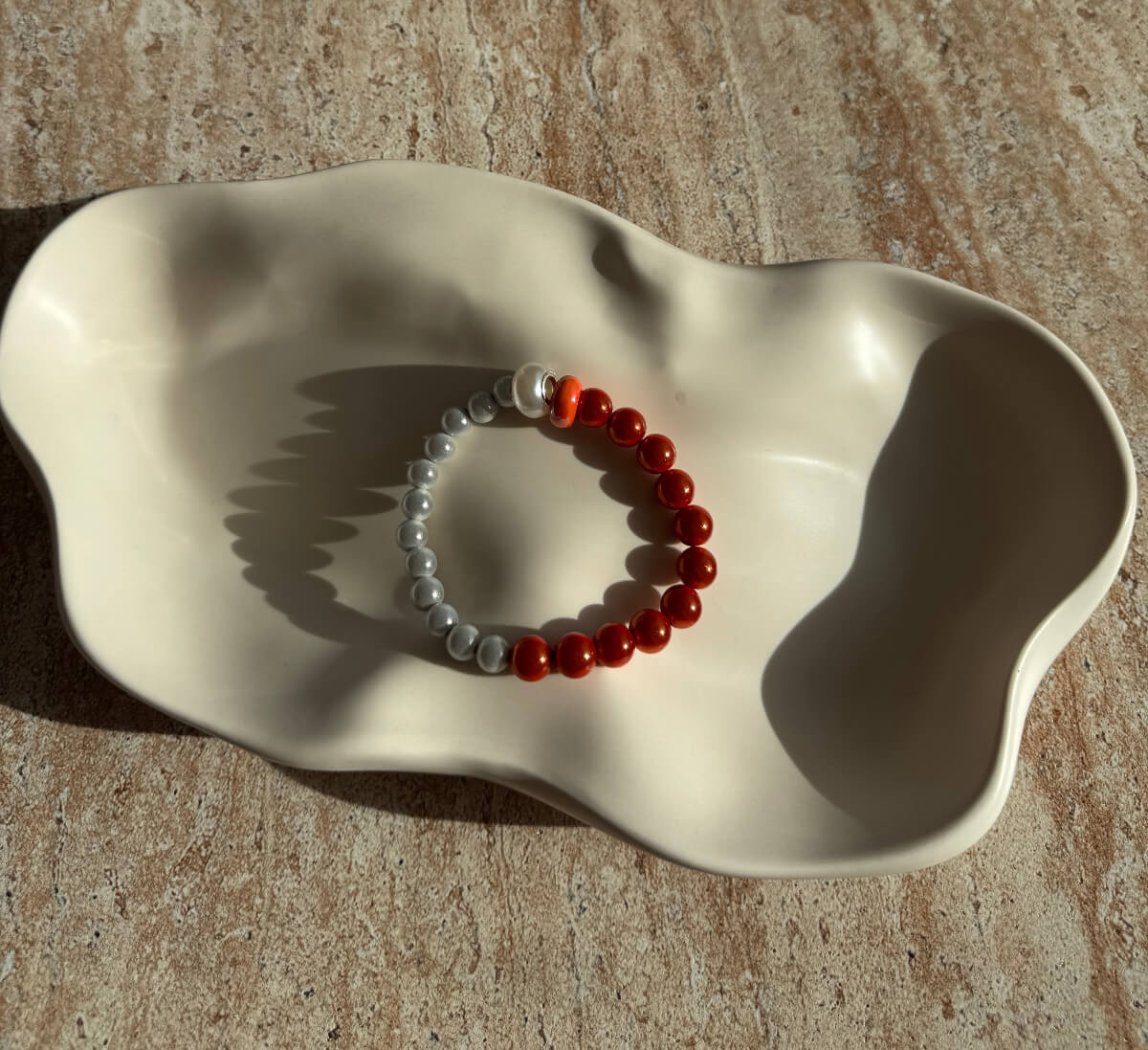 Komplettes Armband mit grauen und orangefarbenen Perlen auf einer cremefarbenen Schale – elegantes, harmonisches Schmuckstück im Sym Aesthetics Stil.