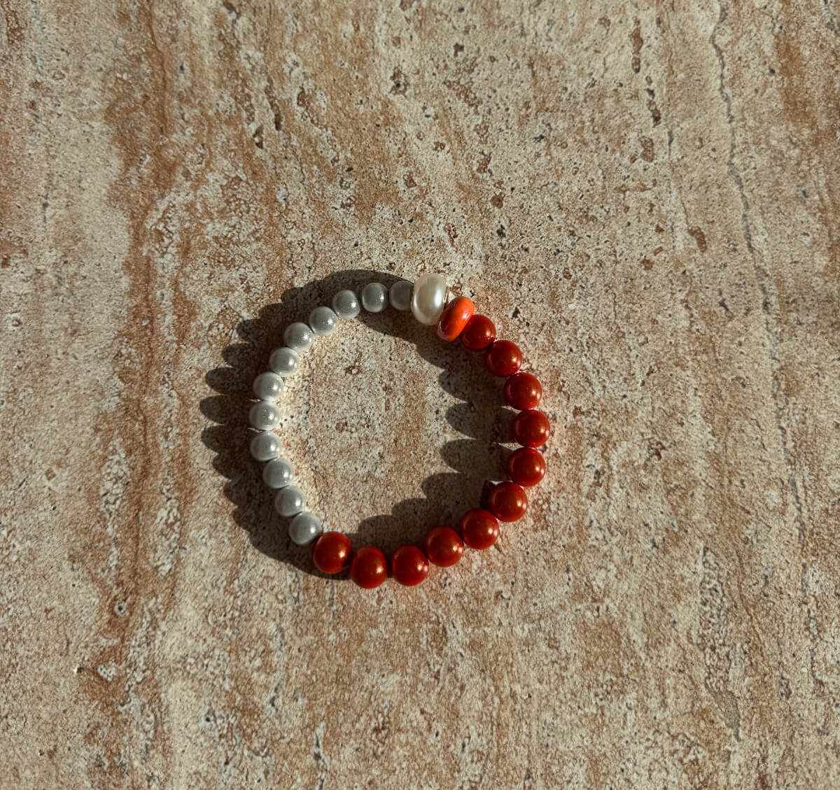 Armband aus grauen und orangefarbenen Perlen liegt auf einer Steinoberfläche im Sonnenlicht – minimalistisches Aesthetic Design.