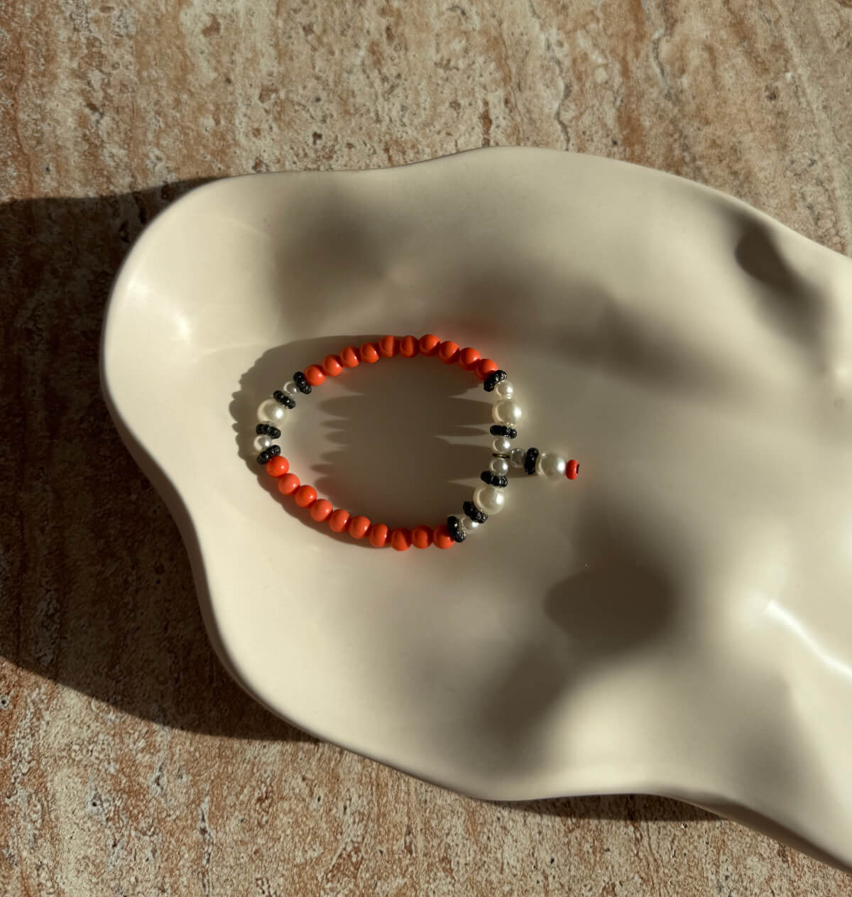 Aesthetic Beads Bracelet in warmen Orangetönen mit weißen und schwarzen Akzenten, liebevoll arrangiert auf einer glatten Keramikschale im Sonnenlicht.