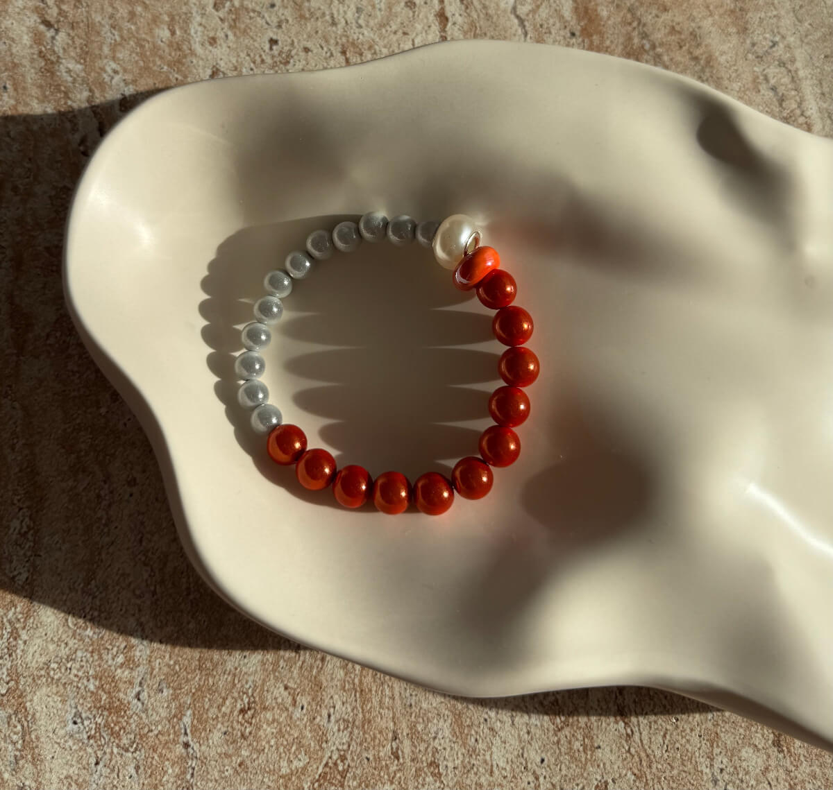 Nahaufnahme eines handgefertigten Armbands mit grauen und orangen Perlen, mittig verziert mit einer weißen und einer bernsteinfarbenen Kugel.