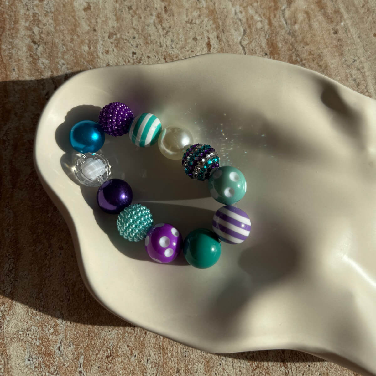 „Nahaufnahme des Mermaid Carnival Armbands in einer weißen Schale mit glitzernden und gestreiften Perlen.“