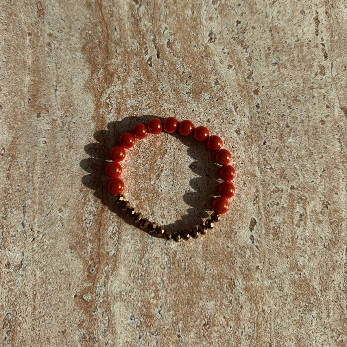Armband aus orangefarbenen und bronzenen Perlen liegt auf einer cremefarbenen Schale im warmen Sonnenlicht.